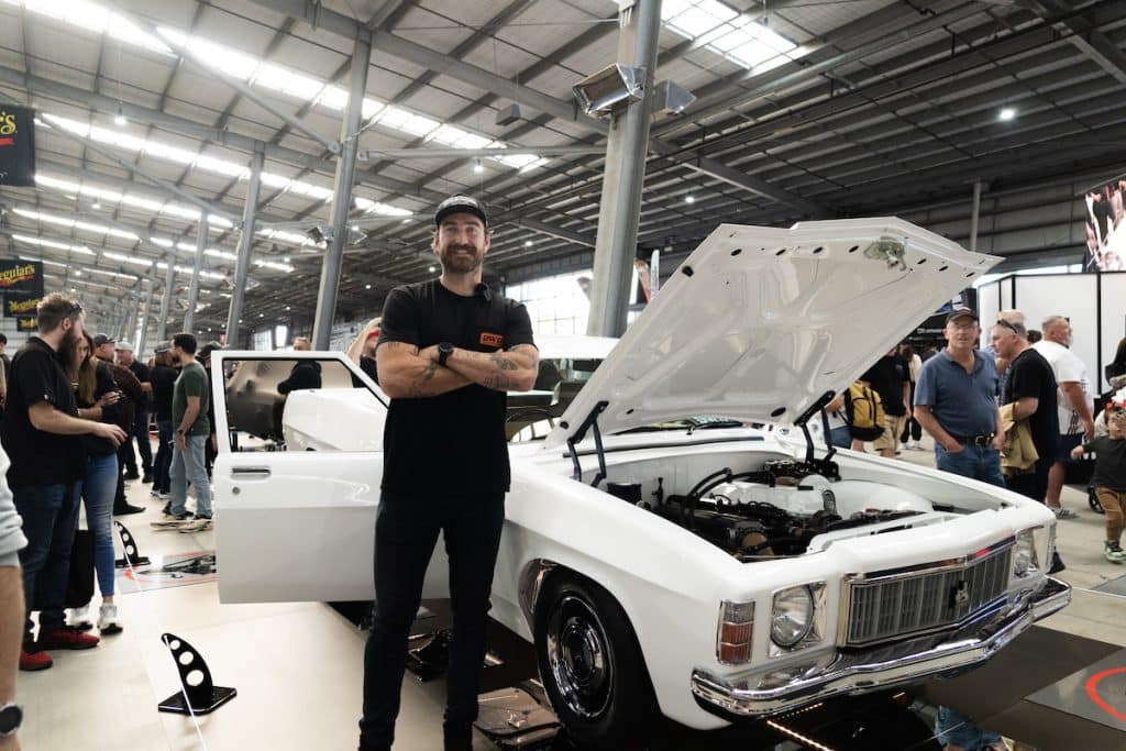 Wide angle shot, indoors at a car show, showing retired Port Adelaide AFL star Chalie Dixon mid frame, standing in front of a modified 1977 HX Kingswoods Ute with the botton up revealing the Billet RB engine with a the RB30 ‘long block’ and high-end turbo