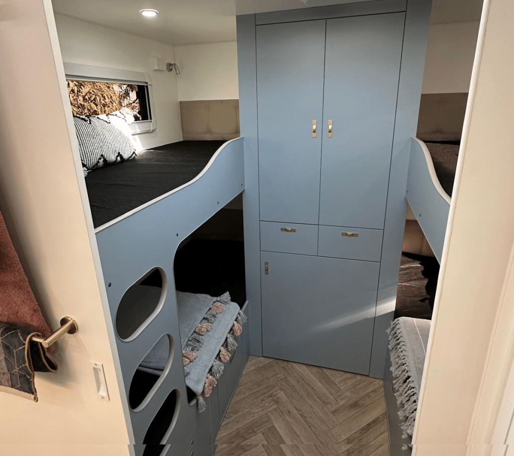 Caravan interior from the mid-ship bathroom showing two sets of bunks separated by a wall to floor cabinets with wardrobes up top and small drawers mid way. A bottom cabinet looks like it conceals a front loading washing machine 