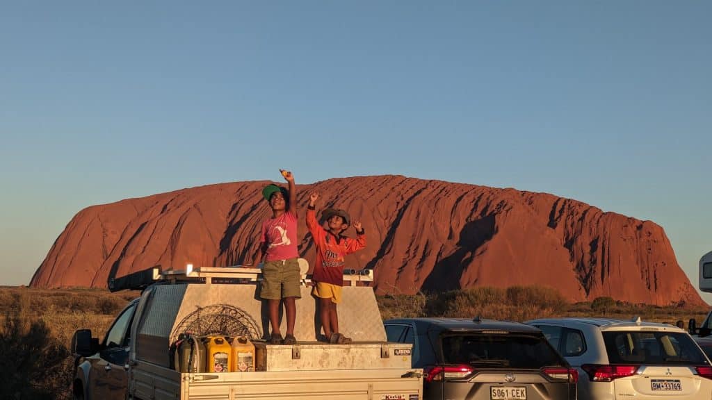 Two boys wearing caps stand atop of the a ute tray toolbox, fitted with jerry cans. They're in front of Uluru trying the optical illusion showing they can 'reach its top;