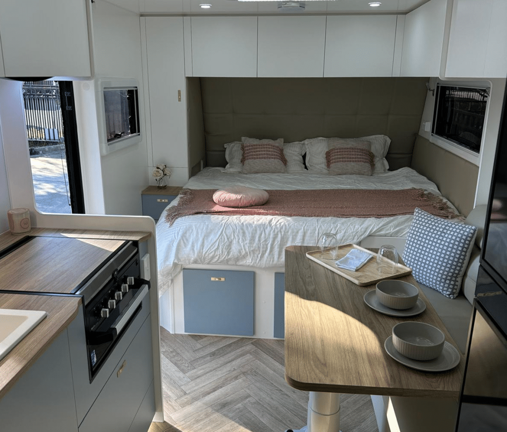 Caravan interior photographed at the kitchen looking towards a king size bed accessible from two sides. Note the bedhead that extends along the back of the bed to the front of the mattress.