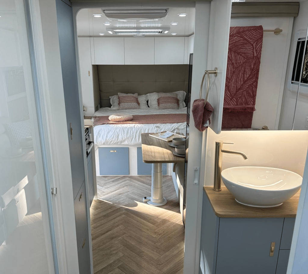 Caravan interior taken from the centre at the ensuite. Note the herringbone pattern on the floor, linen closet in the bathroom and the mirrored medicine cabinent. Below that a china bowl and matt gold faucet are softly lit.